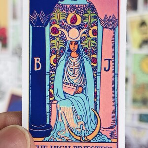 May include: A tarot card sticker with a colorful illustration of the High Priestess. The card is pink, blue, and yellow with a woman in a white robe sitting on a throne. The card has the Roman numeral II and the text "THE HIGH PRIESTESS."
