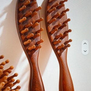 Puede incluir: Dos cepillos de pelo de madera con acabado marrón oscuro. Cada cepillo tiene una cabeza ovalada con numerosas púas de madera redondeadas y un mango largo y curvado. Los cepillos se muestran sobre un fondo neutro, resaltando la veta de la madera.