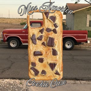 May include: A rectangular phone case featuring a textured, light tan surface resembling a cookie dough base embedded with numerous dark brown chocolate chunks. The case has a cutout for a camera lens and displays the text Miss America Cookie Case.