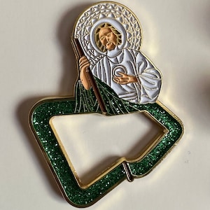 May include: Gold-tone metal pin with a green glitter background. The pin features a religious icon of a man with a halo and a staff. The man is wearing a white robe and holding a heart in his hand.