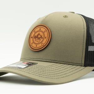 May include: An olive green baseball cap with a black mesh back and a curved brim. A circular tan leather patch is centred on the front, featuring an embossed compass design with mountain peaks and the cardinal directions N, S, E, and W.