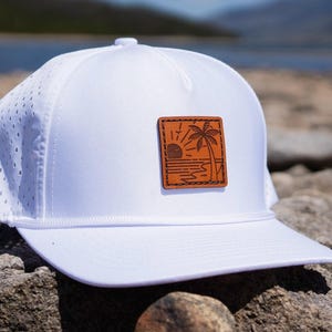 Può includere: Cappellino da baseball bianco in mesh con una toppa in pelle marrone che presenta una grafica di una palma e un tramonto.