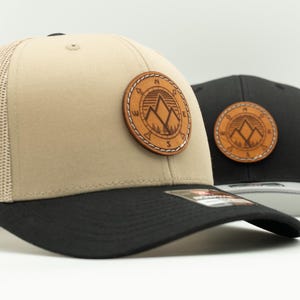 May include: Two trucker hats featuring a tan and a black front panel with matching mesh backs. Each hat has a circular brown leather patch on the front displaying a mountain range graphic with compass directions N, S, E, and W around the border.