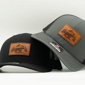May include: A black baseball cap and a gray baseball cap, both featuring a rectangular brown leather patch on the front. Each patch displays a laser-engraved graphic of a bear standing in a forest with mountains in the background.