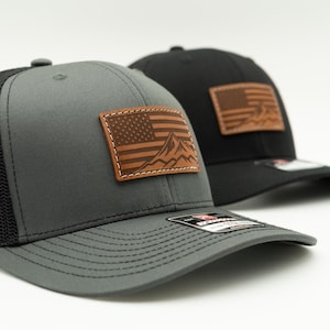 Może przedstawiać: Dwie czapki typu trucker, jedna szara i jedna czarna, ze skórzaną naszywką z amerykańską flagą i górskim wzorem. Szara czapka ma czarną siatkową tylną część. Obie czapki mają zakrzywiony daszek.