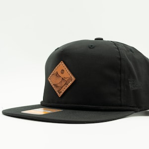 May include: A black baseball cap with a brown leather patch featuring a mountain range and a sun. The patch is in the shape of a diamond.
