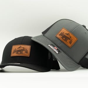 May include: Two trucker caps, one black and one grey, with a brown leather patch showing a bear, trees, and a mountain. The grey cap has a black mesh back and a curved peak.