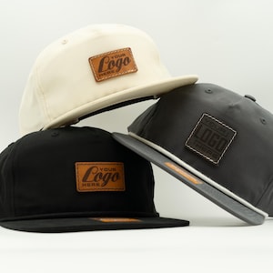 May include: Three baseball caps in cream, black, and charcoal gray. Each cap features a flat brim and a square leather patch with the text "YOUR LOGO HERE". The caps are stacked, showcasing their design and branding.