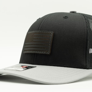 May include: A black and light gray trucker hat featuring a rectangular patch on the front panel with a debossed American flag design. The cap has a solid black front, a light gray brim, and a black mesh back with an adjustable snapback closure.
