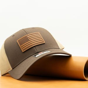 May include: A brown trucker hat with a tan mesh back and a leather patch featuring the American flag. The hat is resting on a brown surface.