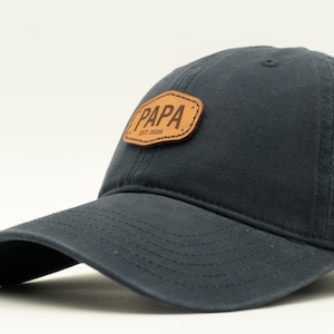 May include: A dark charcoal grey baseball cap featuring a tan leather-like patch on the front panel. The patch is rectangular with rounded corners and displays the text "PAPA" in a bold font, with "EST. 2020" printed in smaller text underneath.