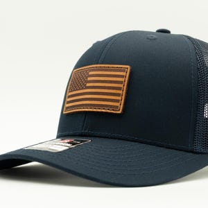 May include: A navy blue trucker hat featuring a rectangular brown leather patch on the front panel. The patch is embossed with the design of the American flag. The hat has a structured front, a curved brim, and a breathable mesh back panel.