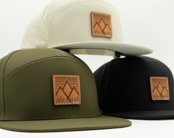 May include: Three five-panel caps in olive green, cream, and black. Each hat features a square brown leather patch on the front panel, embossed with a graphic of mountain peaks and pine trees, and includes perforated side panels for ventilation.