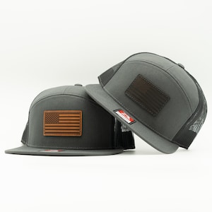 May include: Two gray trucker hats with a brown leather American flag patch. The hats have a mesh back and a flat brim. One hat is slightly tilted, showing the patch and the brim.