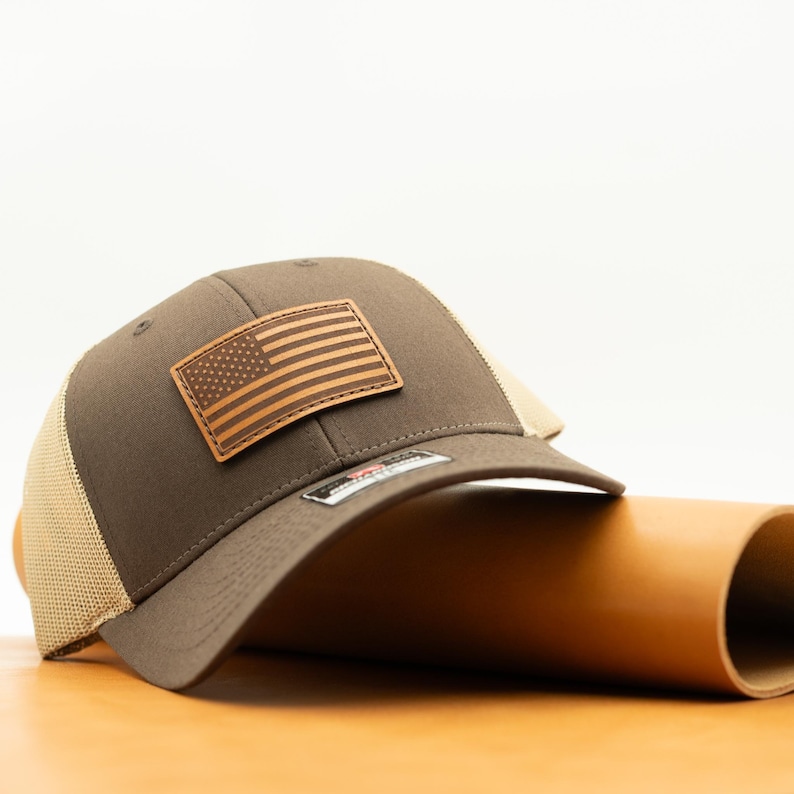 May include: A brown trucker hat with a tan mesh back and a leather patch featuring the American flag. The hat is resting on a brown surface.