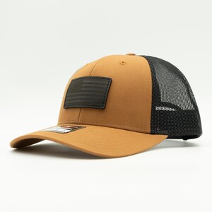 May include: A brown trucker hat with a black mesh back and a curved brim. The hat features a black patch with an American flag design on the front.