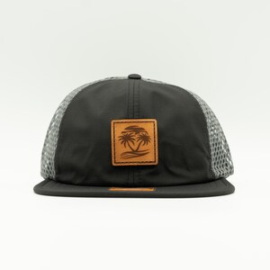 May include: A black and gray mesh baseball cap with a brown leather patch featuring a palm tree design.