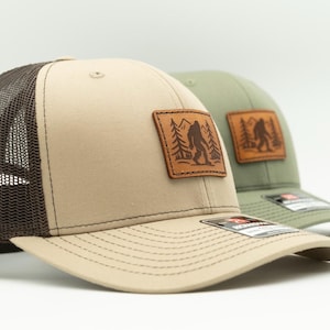 May include: Two baseball caps with a Bigfoot patch. One cap is tan with a brown mesh back, the other is olive green. Both have a leather patch with a Bigfoot silhouette in a forest.