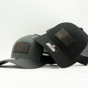 May include: Two baseball caps, one charcoal gray and one black, are displayed. Each cap features a brown leather patch with an American flag design. The black cap has a mesh back. Both caps have a curved brim.