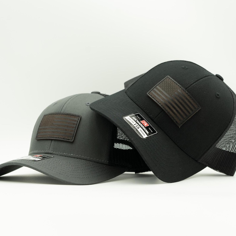 May include: Two baseball caps, one charcoal gray and one black, are displayed. Each cap features a brown leather patch with an American flag design. The black cap has a mesh back. Both caps have a curved brim.