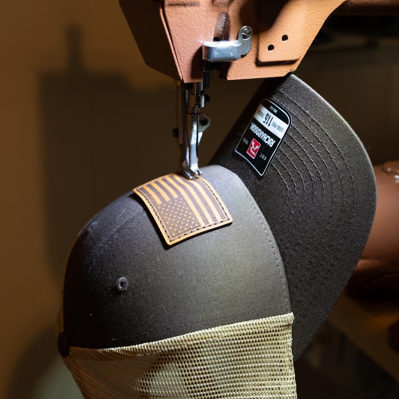 May include: A brown and tan trucker hat with a leather American flag patch is being sewn by a machine. The hat has a mesh back and a curved brim. A black and white label with the brand name "RICHARDSON" is visible.