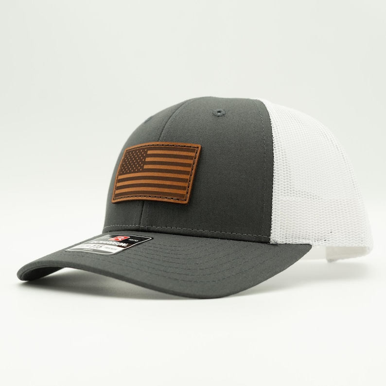 May include: A charcoal gray baseball cap with a white mesh back. The cap features a brown leather patch with an American flag design. The brim is curved, and the cap has a structured fit.