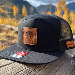 Op de afbeelding: Zwarte en bruine truckercap met een platte klep en een leren patch met een berglandschap en de tekst "FLAT BILL SNAPBACK".