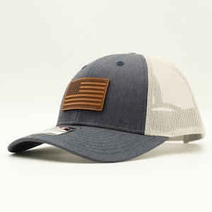 May include: A navy blue trucker hat with a tan mesh back and a curved brim. The hat features a brown leather patch with an American flag design. The hat is a classic style, suitable for outdoor activities.