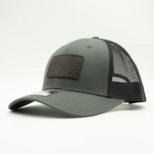 May include: Gray trucker hat with a black mesh back and a curved brim. A brown leather patch on the front displays an American flag design. The hat has a structured fit and a snapback closure.