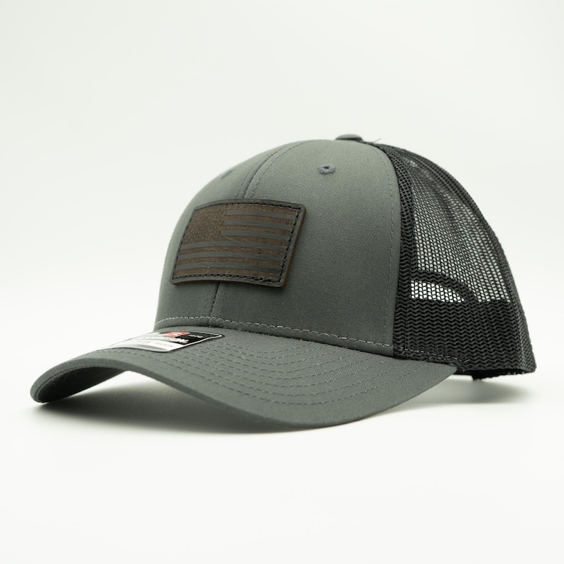May include: Gray trucker hat with a black mesh back and a curved brim. A brown leather patch on the front displays an American flag design. The hat has a structured fit and a snapback closure.
