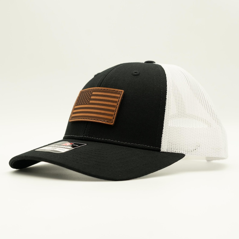 May include: Black and white trucker hat with a brown leather American flag patch. The hat features a curved brim and a mesh back for breathability. A stylish accessory for everyday wear.