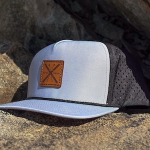 May include: A light blue and black mesh trucker hat with a brown leather patch featuring a crossed ski design and the text "OUTLAW" and "LYNXES".