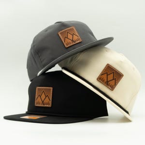 May include: Three baseball caps in gray, black, and cream colors are stacked. Each cap features a square, brown leather patch with a mountain and tree design. The caps have a flat brim and a rope detail.