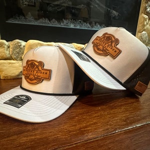 May include: Two white and black trucker hats with a leather patch featuring the text "18 Pucks Given Podcast" and hockey sticks. The hats have a curved brim and a black rope detail. One hat has a black mesh back and the other has a white mesh back.