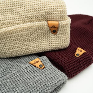 May include: Three knitted beanies in different colors: beige, gray, and burgundy. Each beanie has a small leather patch with a logo on the side.