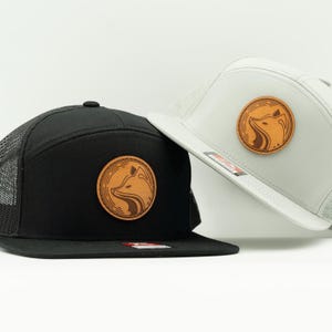 Puede incluir: Dos gorras de camionero, una negra y otra gris, con un parche con el logotipo de un zorro. La gorra negra tiene la parte trasera de malla negra y una visera plana. La gorra gris tiene la parte trasera de malla gris y una visera plana. Ambas gorras tienen un parche de cuero marrón con un diseño de zorro.