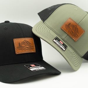 May include: Two trucker caps are shown, one black and one olive green. Each cap has a leather patch with a mountain and tree design. The black cap has a black mesh back, while the olive cap has a grey mesh back. Both caps have the "RICHARDSON 112" label.