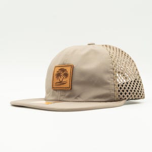 May include: A beige mesh and canvas baseball cap with a brown leather patch featuring a palm tree design.