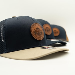 Puede incluir: Una gorra tipo trucker azul marino con visera plana color canela y parte trasera de malla oscura. El frente presenta un parche circular de cuero marrón grabado con un diseño de montañas, bosque y sol. Varias gorras idénticas están alineadas.