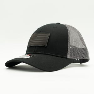 May include: Black trucker hat with a gray mesh back and a curved brim. The hat features a dark gray American flag patch on the front. The hat is a classic style, suitable for casual wear.