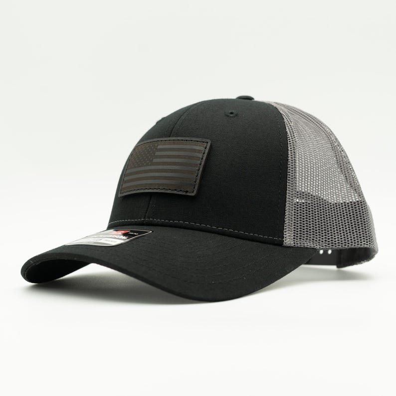 May include: Black trucker hat with a gray mesh back and a curved brim. The hat features a dark gray American flag patch on the front. The hat is a classic style, suitable for casual wear.