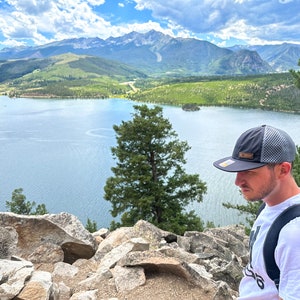 Lifestyle photo of man wearing Richardson 935 Active Mountain performance hat while looking over alpine lake and mountains, showcasing water resistant leather patch outdoors hat in use