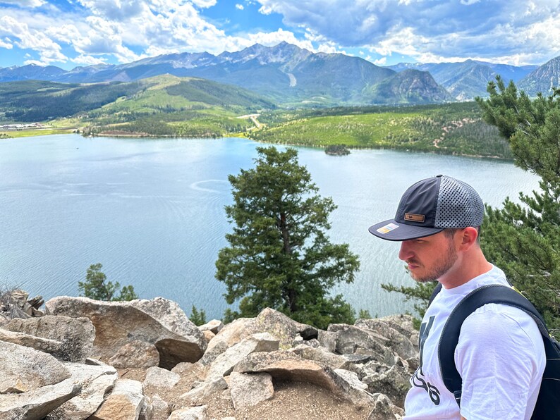 Lifestyle photo of man wearing Richardson 935 Active Mountain performance hat while looking over alpine lake and mountains, showcasing water resistant leather patch outdoors hat in use