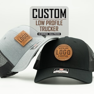 May include: A light gray and a black trucker hat, each featuring a brown leather patch with the text 'YOUR LOGO HERE'. The text 'CUSTOM LOW PROFILE TRUCKER' is displayed above the hats. The black hat has a Richardson 115 sticker on the brim.