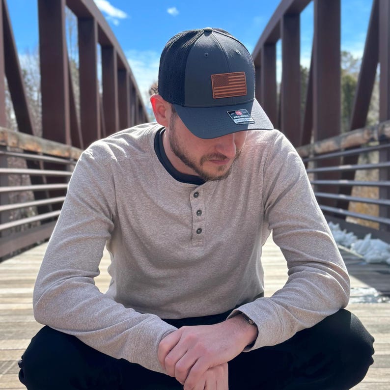 May include: A dark gray trucker hat with a black mesh back and a brown leather patch featuring a red, white, and blue American flag. The hat is worn by a person wearing a light gray long-sleeve shirt.