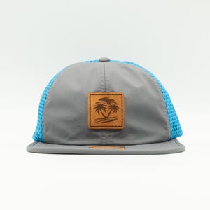 May include: A gray and blue mesh trucker hat with a brown leather patch featuring a palm tree design.