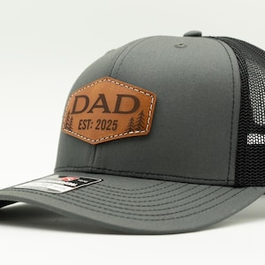 May include: Gray trucker hat with a black mesh back and a curved brim. The front features a brown leather patch with the word "DAD" and "EST: 2025" in a serif font, along with tree illustrations. The hat has a snapback closure.