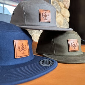 May include: Three 5-panel caps in different colours: grey, navy blue, and olive green. Each cap features a brown leather patch with a stylised image of three pine trees.