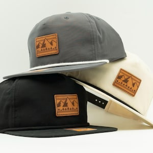 May include: Three baseball caps in gray, black, and cream colors. Each cap features a flat brim and a leather patch with a mountain and tree design. A white rope detail accents the gray cap.