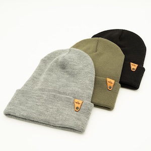 May include: Three knit beanies in gray, olive green, and black. Each beanie has a leather patch with a logo on the front.